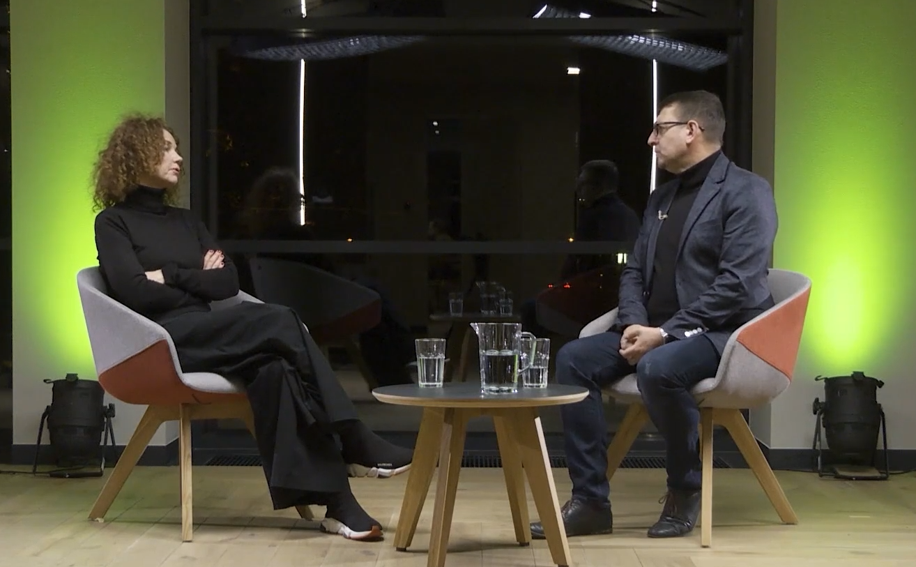 Kobieta i mężczyzna siedzą na fotelach po dwóch stronach małego stolika na tle dużego okna i rozmawiają